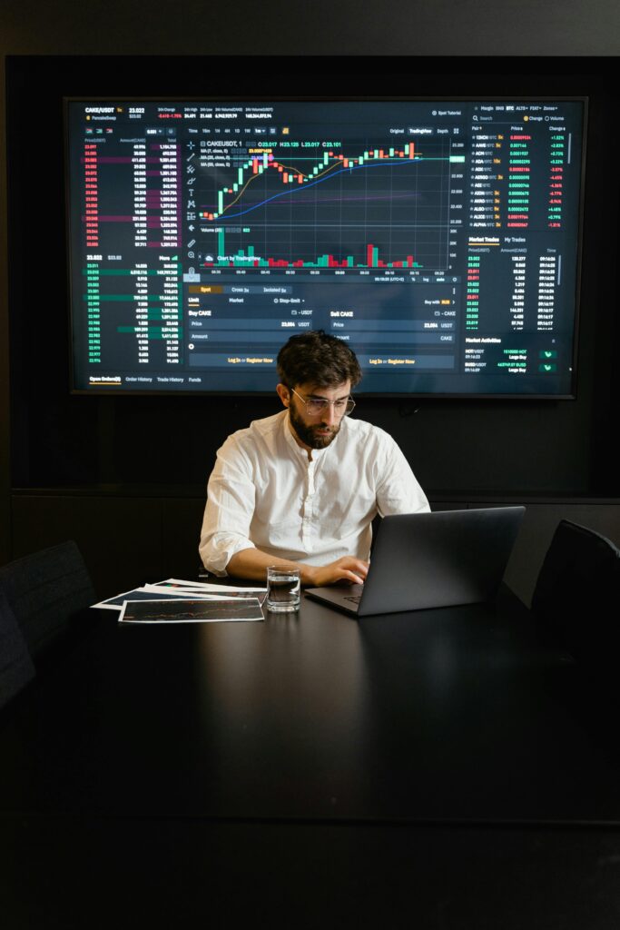 A focused man analyzing stock market trends on a laptop in an office setting with charts displayed.